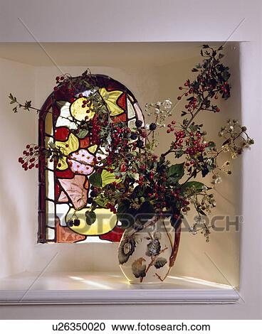 Stock Image - Close-up of autumn berries in vase in alcove with small stained glass window. Fotosearch