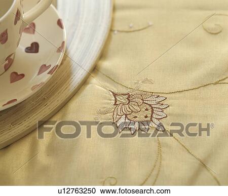 Stock Image - Close-up of heart-patterned cup on cream silk embroidered quilt. Fotosearch
