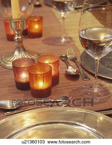 Stock Image - Close-up of lighted candles in orange jars on table with wineglasses and metal plates. Fotosearch