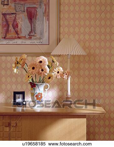 Stock Photography - Close-up of pink gerberas in vase on shelf with lamp in front of peach checked wallpaper. Fotosearch