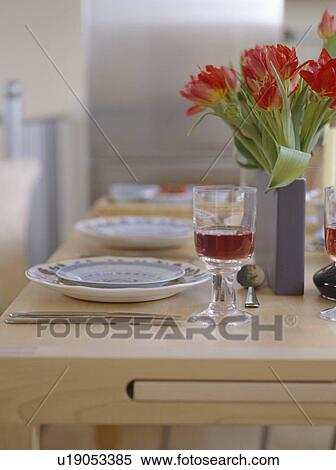Stock Photography - Close-up of red tulips in grey vase on pale wood table with place settings and glass of red wine. Fotosearch