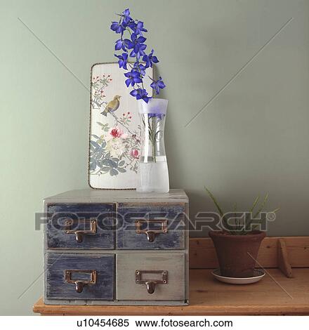 Stock Photography - Close-up of small Chinese picture and single blue delphinium flower in glass vase on set of small filing drawers. Fotosearch