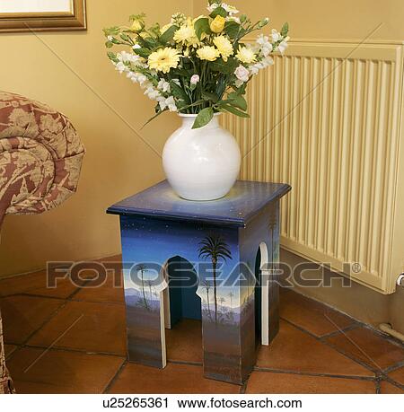 Stock Image - Close-up of yellow flowers in white vase on small wooden table with painted trompe l'oeil effect. Fotosearch