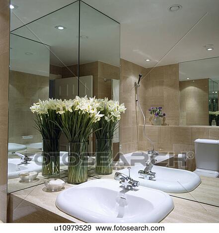 Stock Photo - Closeup of mirrored bathroom walls, white basin and yellow flowers in glass vase. Fotosearch