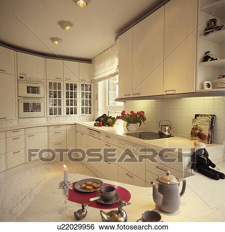 Stock Photograph - Coffee pot on curved peninsular unit in modern white kitchen with ceiling spotlights and recessed lighting below cupboards. Fotosearch