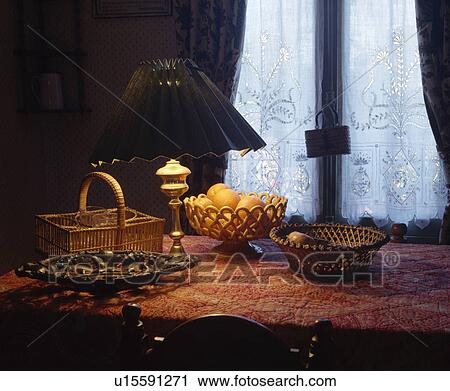 Stock Image - Collection of cane and ceramic baskets with lighted lamp on table in front of lace curtain. Fotosearch