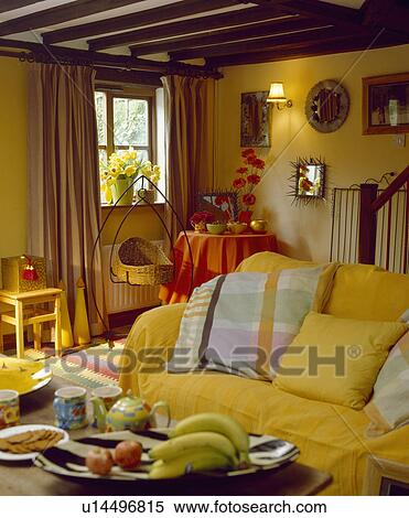 Stock Photography - Colourful cushions and yellow throw on sofa in yellow cottage livingroom with rocking wicker cradle on metal frame. Fotosearch