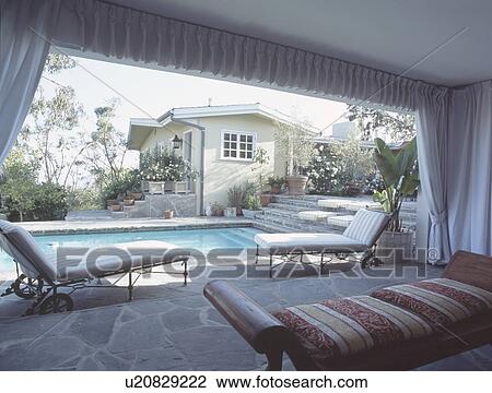 Stock Image - Comfortable loungers on stone flooring in veranda living room beside swimming pool in Los Angeles. Fotosearch