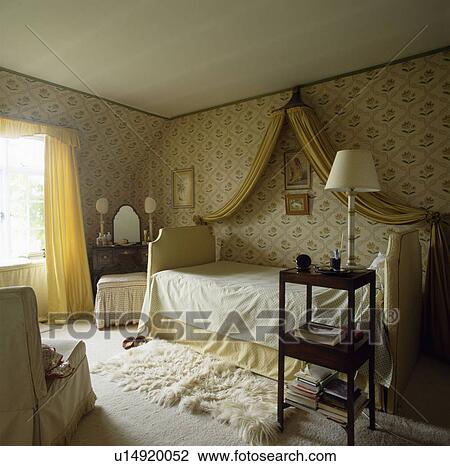 Coronet With Yellow Drapes Above Bed Placed Sideways In Bedroom With Patterned Wallpaper And Sheepskin Rug Stock Image