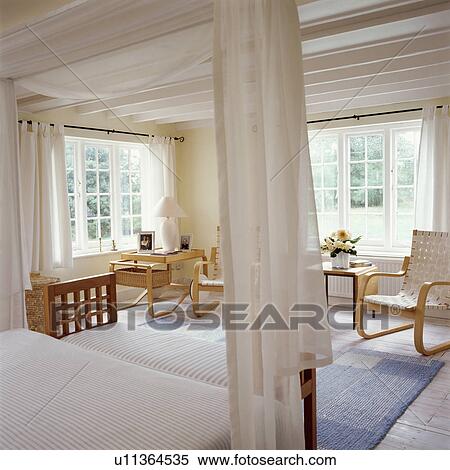 Stock Photography - Cream voile curtains on modern fourposter bed in white country bedroom with large windows. Fotosearch