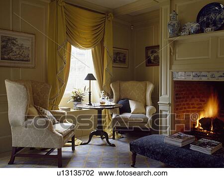 Stock Image - Cream wingchairs in cream panelled livingroom with yellow swagged curtains and lighted fire in fireplace. Fotosearch