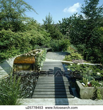 Stock Image - Decking bridge beside waterfall and pond with waterlilies in corner of garden. Fotosearch