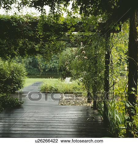 Stock Image - Decking path through a rose covered pergola to a lawn, seating area and still water.. Fotosearch