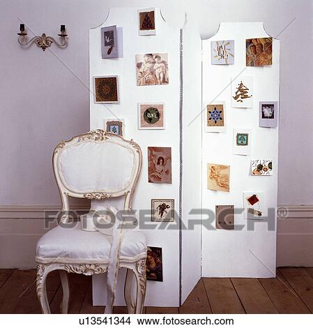 Picture - Detail shot of traditional dining chair upholstered in a white fabric in front of a white screen decorated with postcards. Fotosearch