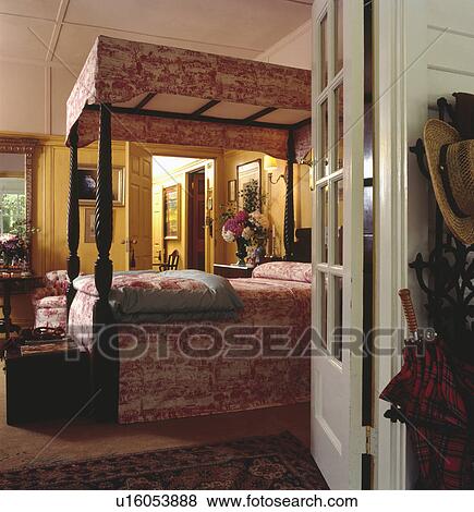 Stock Photo - Door open to country bedroom with red & white "Toile de Jouy" pelmet and bedcover on antique fourposter bed. Fotosearch