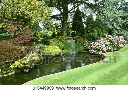 Stock Photo - Fountains in large curved pond in formal country garden with pink hydrangeas. Fotosearch