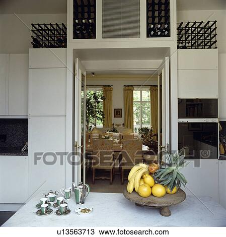 Stock Image - Fruit and antique coffee set on table in modern white kitchen with wine storage above open double doors to dining room. Fotosearch