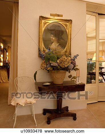 Stock Photo - Gilt-framed Edwardian picture above pot of dried flowers on antique mahogany pedestal table beside white chair in hall. Fotosearch
