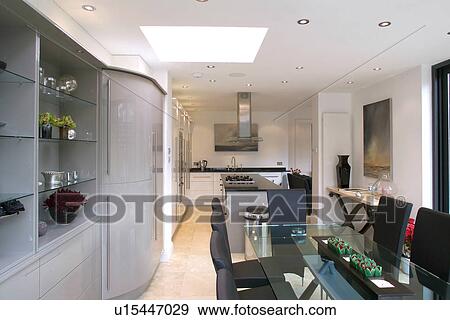 Stock Photo - Glass table and upholstered black chairs in modern openplan white kitchen and dining room with fitted shelving unit. Fotosearch