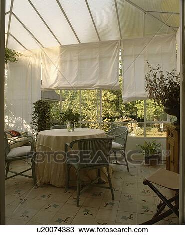 Stock Image - Green wicker chairs circular table with cloth in conservatory dining room with white blinds . Fotosearch