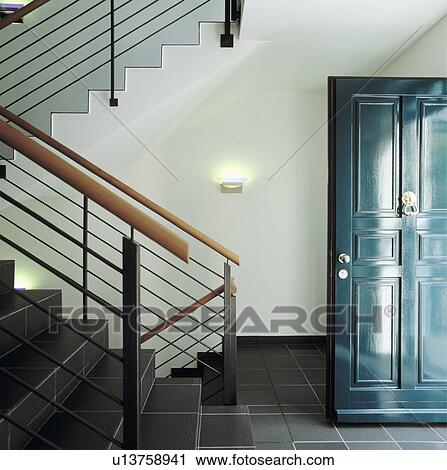 Stock Image - Grey floor tiles in modern hall with blue open front door and wood and metal banniesters on staircase. Fotosearch