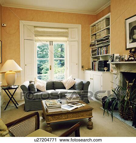 Grey sofa in front of French windows with shutters in livingroom with bookshelves and carved wood coffee table View Large Photo Image Stock Image - Grey sofa in front of French windows with shutters in livingroom with bookshelves and carved wood coffee table. Fotosearch