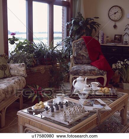Stock Image - Houseplants in front of window with chess game and tray set for tea with teapot and cup on low table in front of armchair and sofa. Fotosearch