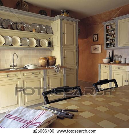 Stock Image - Knives on PVC tablecloth in country kitchen with plates on shelves above sink in fitted cream cupboard. Fotosearch