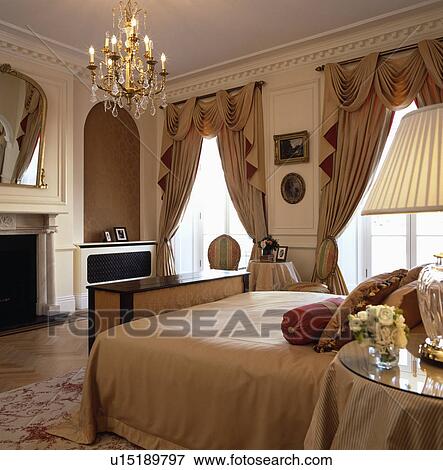 Lighted antique glass chandelier above bed bed in opulent bedroom with swagged and tailed curtains View Large Photo Image Stock Photo - Lighted antique glass chandelier above bed bed in opulent bedroom with swagged and tailed curtains. Fotosearch