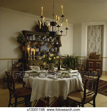 Lighted candle chandelier above place settings and white cloth on circular table in dining room with antique dresser View Large Photo Image Stock Photograph - Lighted candle chandelier above place settings and white cloth on circular table in dining room with antique dresser. Fotosearch