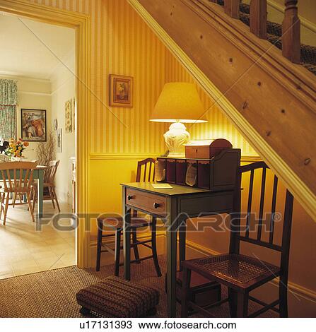 Stock Image - Lighted lamp on antique desk with under the stairs in traditional hall with yellow striped wallpaper. Fotosearch