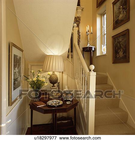 Stock Photo - Lighted lamp on antique table beside stairs with beige carpet in small neutral hall. Fotosearch