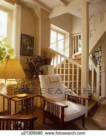 Stock Image - Lighted lamp on table beside Edwardian chair with white cushions and wooden arms beside stairs in white country hall. Fotosearch