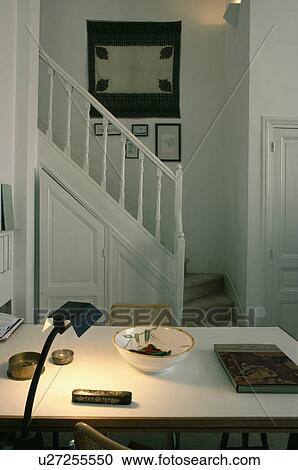 Stock Image - Lighted lamp on table in in small white hall study with picture above staircase. Fotosearch