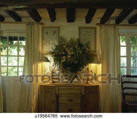 Lighted lamps and flower arrangement on cupboard between windows in white cottage living room View Large Photo Image Stock Photography - Lighted lamps and flower arrangement on cupboard between windows in white cottage living room. Fotosearch