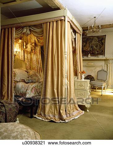 Lighting Inside Opulent Gold Curtains On Fourposter Bed With Floral Cushions And Quilt In Large Country Bedroom Stock Image
