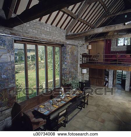 Long table in front of large windows in dining room in barn conversion with stone floor and apex ceiling View Large Photo Image Stock Photo - Long table in front of large windows in dining room in barn conversion with stone floor and apex ceiling. Fotosearch