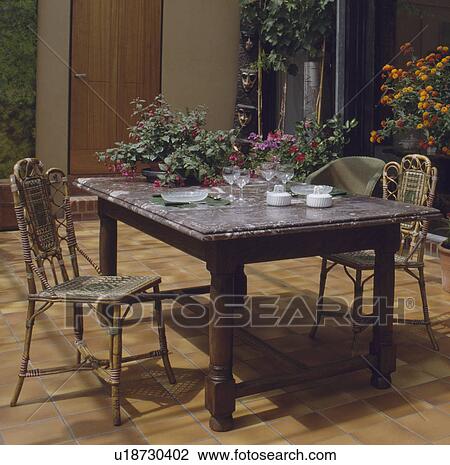 Stock Image - Marble-topped table and cane chairs in conservatory dining room with tiled floor. Fotosearch