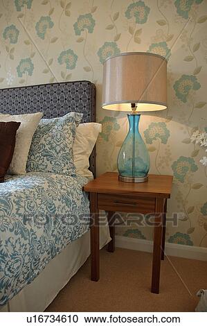 Stock Image - Modern blue flower-patterned wallpaper in bedroom with blue glass lamp on wood bedside table and blue and white bed linen. Fotosearch