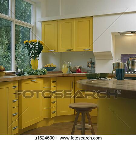 Stock Image - Modern kitchen with yellow cupboards and units and wooden stool at central breakfast bar. Fotosearch