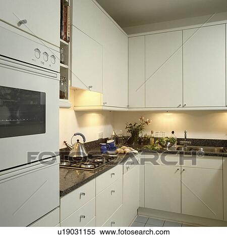 Stock Photography - Modern white kitchen with recessed lighting below fitted cupboards and Alessi kettle on hob in granite worktop. Fotosearch