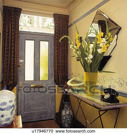 Stock Image - Old black telephone and gladioli in yellow vase on console table in pastel yellow hall with curtains and grey door. Fotosearch