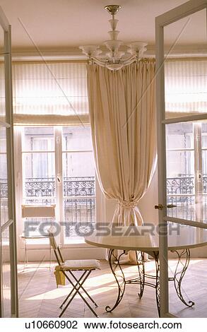 Stock Image - Open doors to circular metal and wood table in diningroom with cream curtain between French windows. Fotosearch