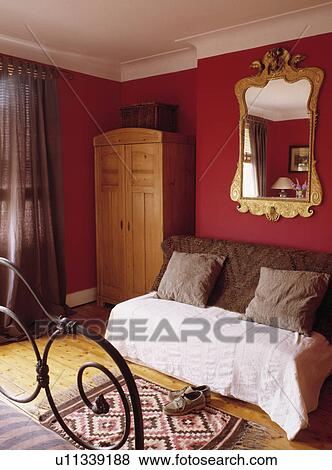 Ornate gilt mirror above couch with brown cushions and cream throw in red bedroom with pine wardrobe View Large Photo Image Stock Photo - Ornate gilt mirror above couch with brown cushions and cream throw in red bedroom with pine wardrobe. Fotosearch