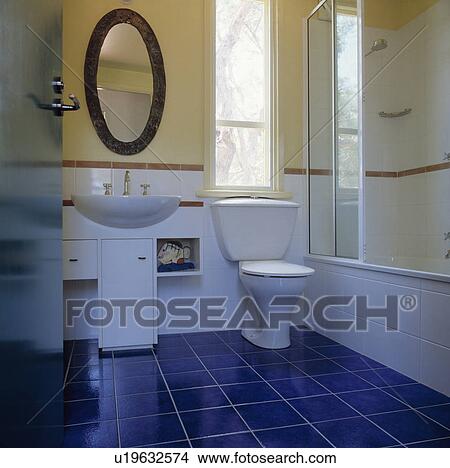 Oval mirror above basin in fitted vanity unit beside toilet in modern bathroom with blue floor tiles View Large Photo Image Picture - Oval mirror above basin in fitted vanity unit beside toilet in modern bathroom with blue floor tiles. Fotosearch