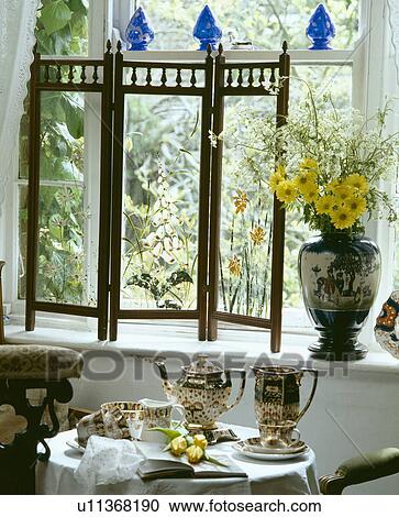 Stock Image - Painted Victorian glass screen on windowsill above table set for tea with teapot and cups. Fotosearch