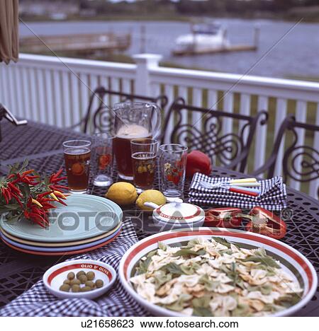Stock Image - Pastel-coloured plates and blue check napkins on table set for lunch with pasta dish and jug of Pimms with glasses. Fotosearch