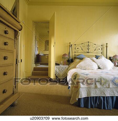 Patchwork quilt and lace cushions on wrought iron bed in yellow country bedroom with door open to ensuite bathroom View Large Photo Image Stock Photo - Patchwork quilt and lace cushions on wrought iron bed in yellow country bedroom with door open to ensuite bathroom. Fotosearch