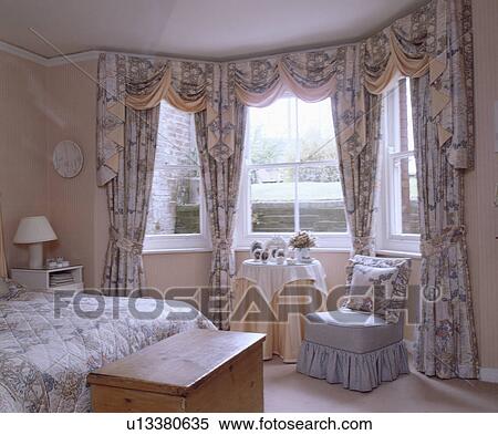 Patterned peach swagged and tailed curtains at bay window above blue chair and small table in traditional bedroom View Large Photo Image Stock Photography - Patterned peach swagged and tailed curtains at bay window above blue chair and small table in traditional bedroom. Fotosearch