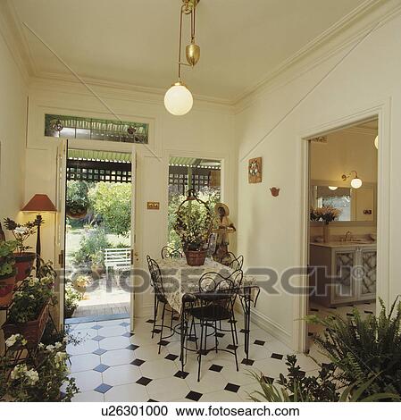 Stock Image - Pendant globe light in dining room with metal chairs and chequerboard tiled floor and open door to garden. Fotosearch